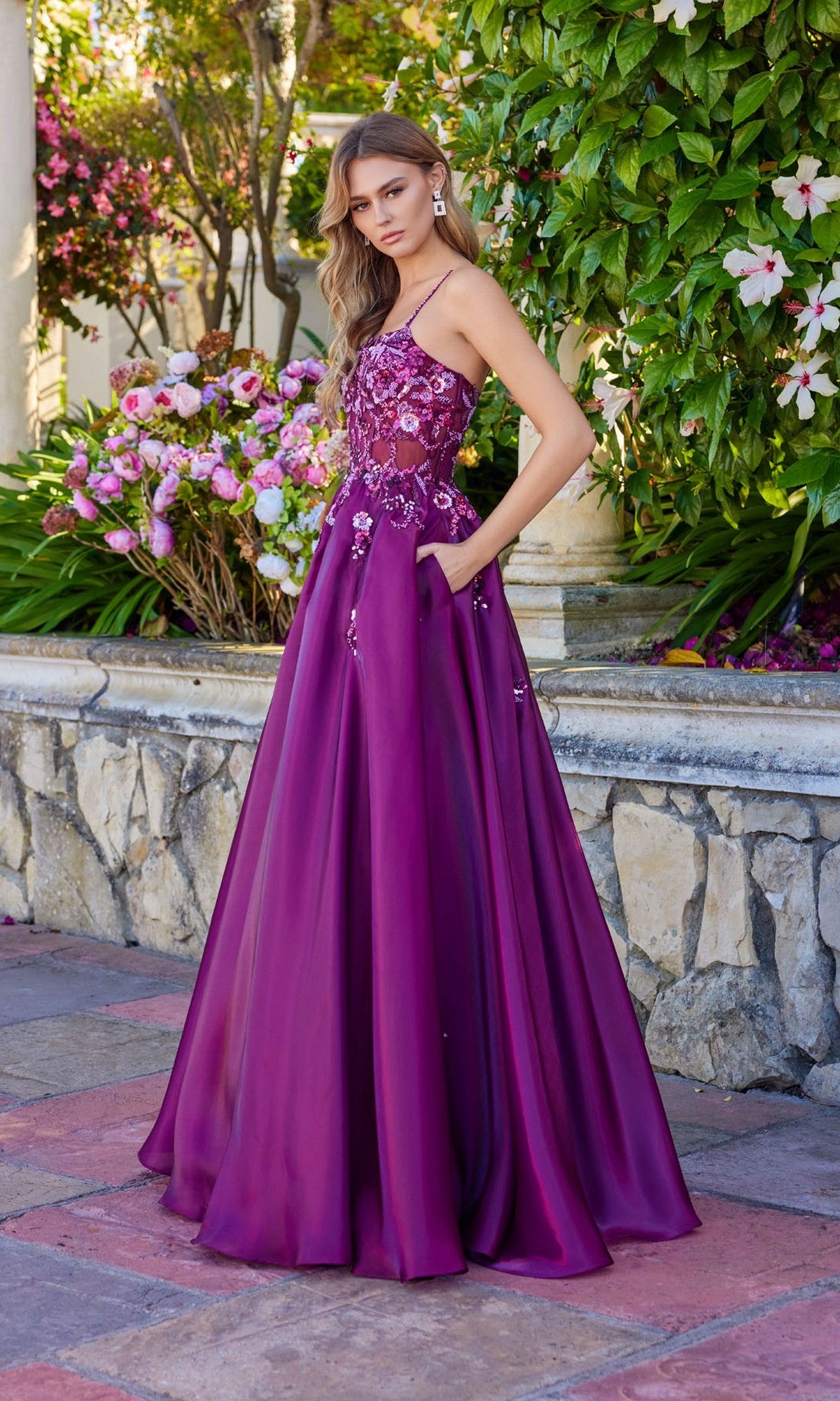 A woman stands outdoors among greenery and pink flowers, wearing the Juliet JT2617U long magenta sleeveless A-line prom dress with floral embellishments.